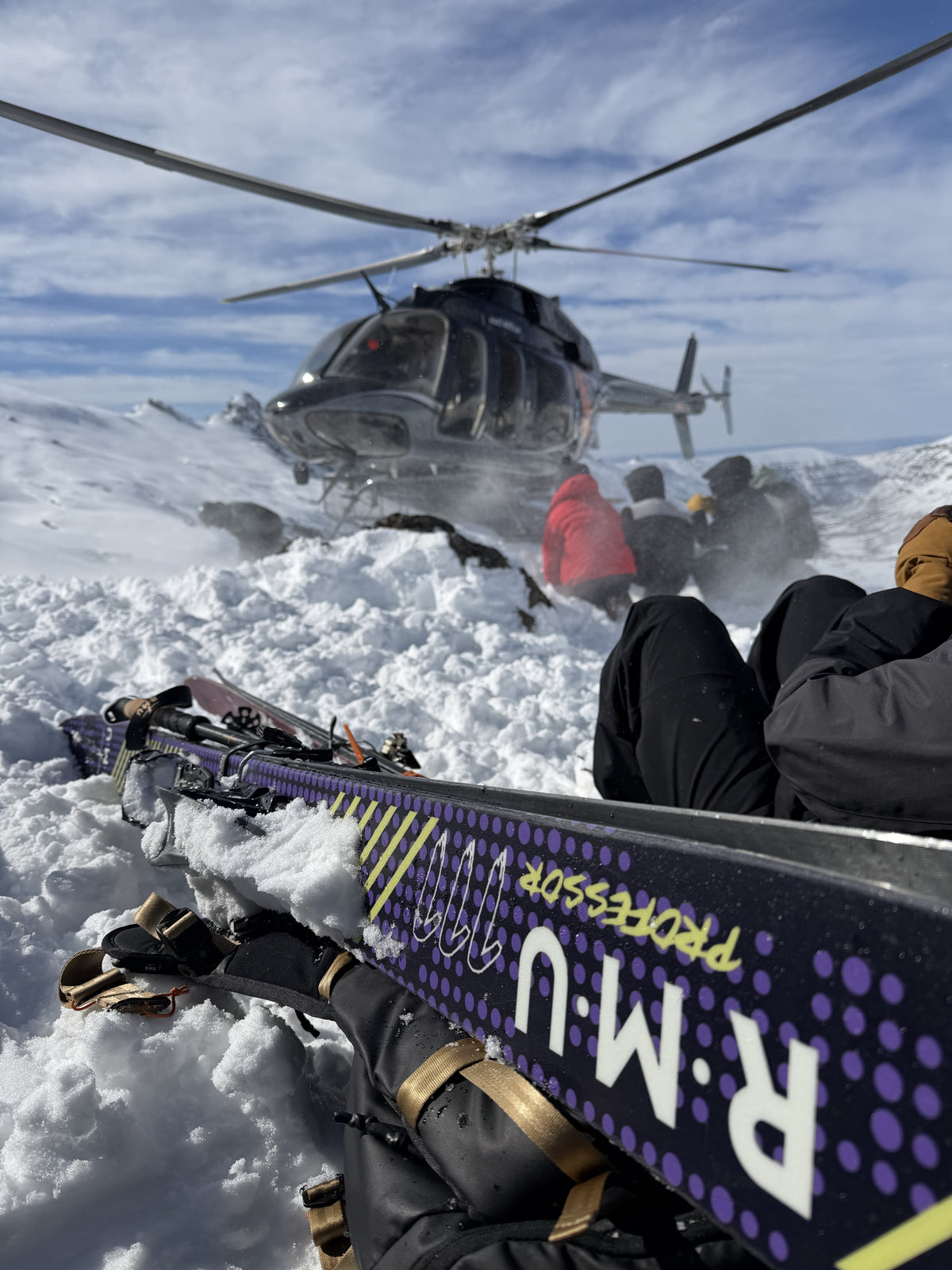 RMU Professor 111 ski in the foreground with a helicopter and crew on the mountain behind